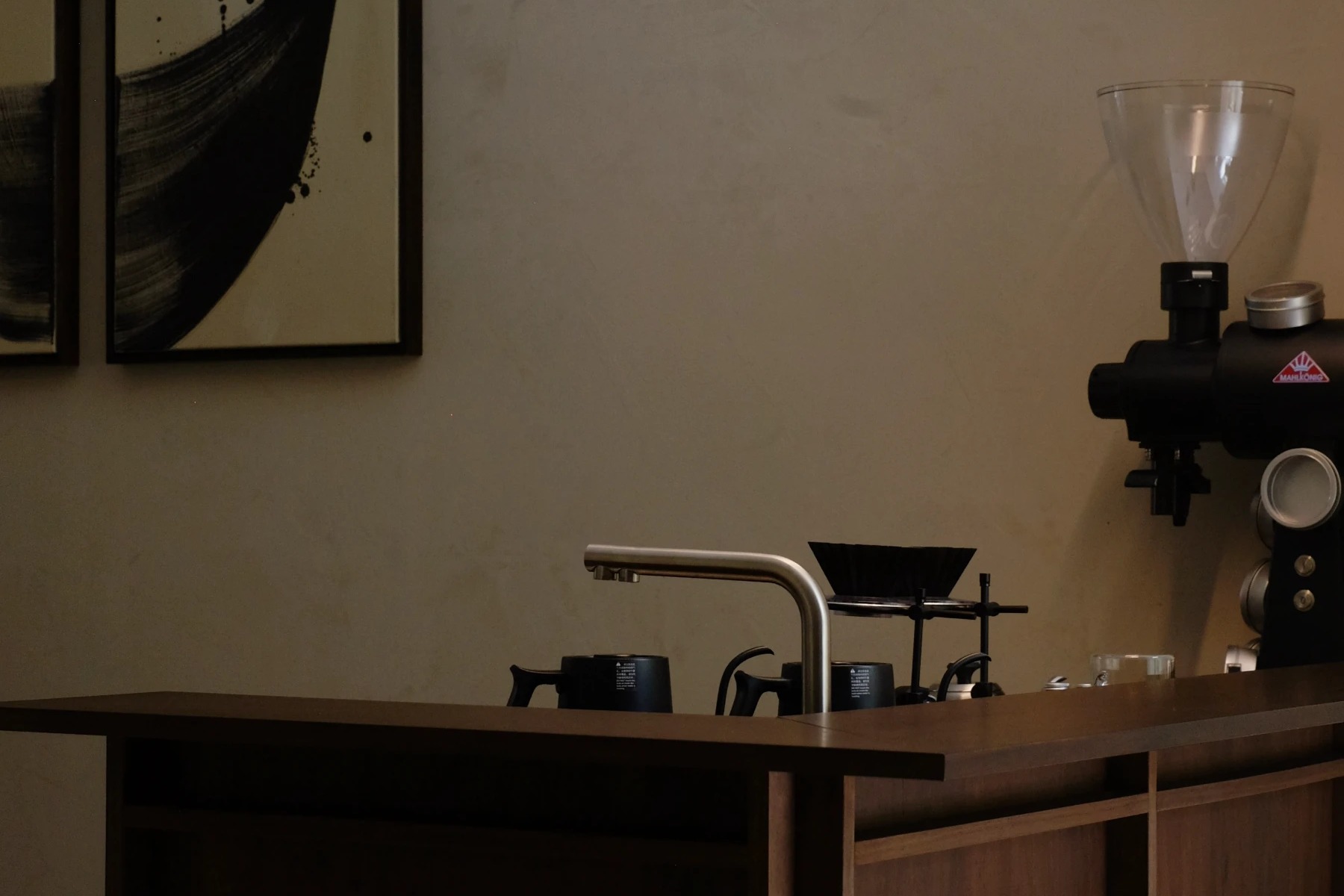 Haku's brew bar with a Mahlkonig grinder and pour over gear beneath framed ink brush artwork, a calm dimly lit interior