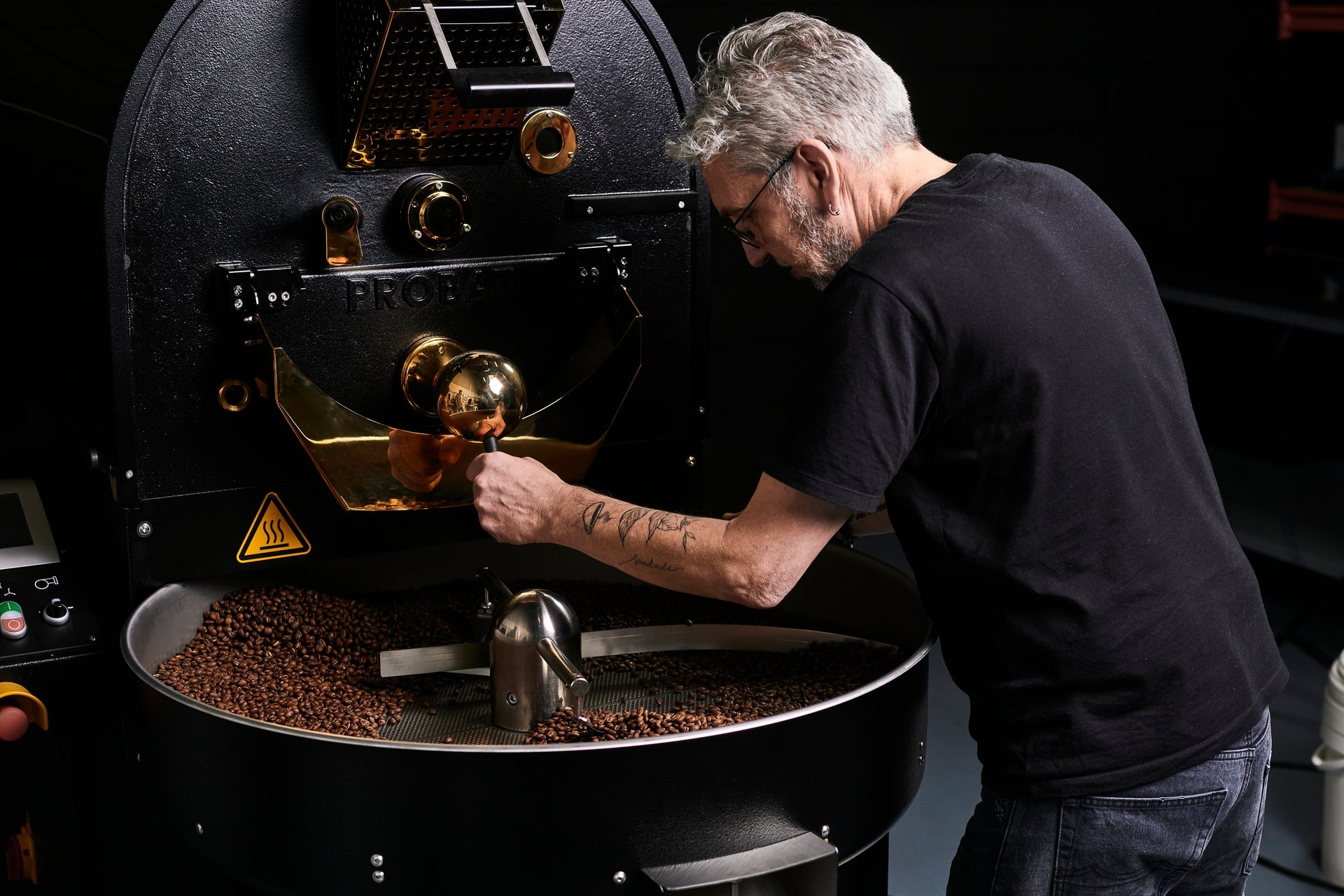 A roaster at Monks Coffee Roasters checks a batch of beans cooling in the tray of a black and brass Probat drum roaster