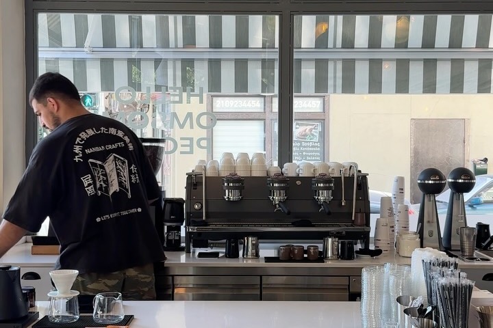 A barista working behind the counter at Omsom, with the espresso machine, pour-over gear and the striped-awning street window behind him