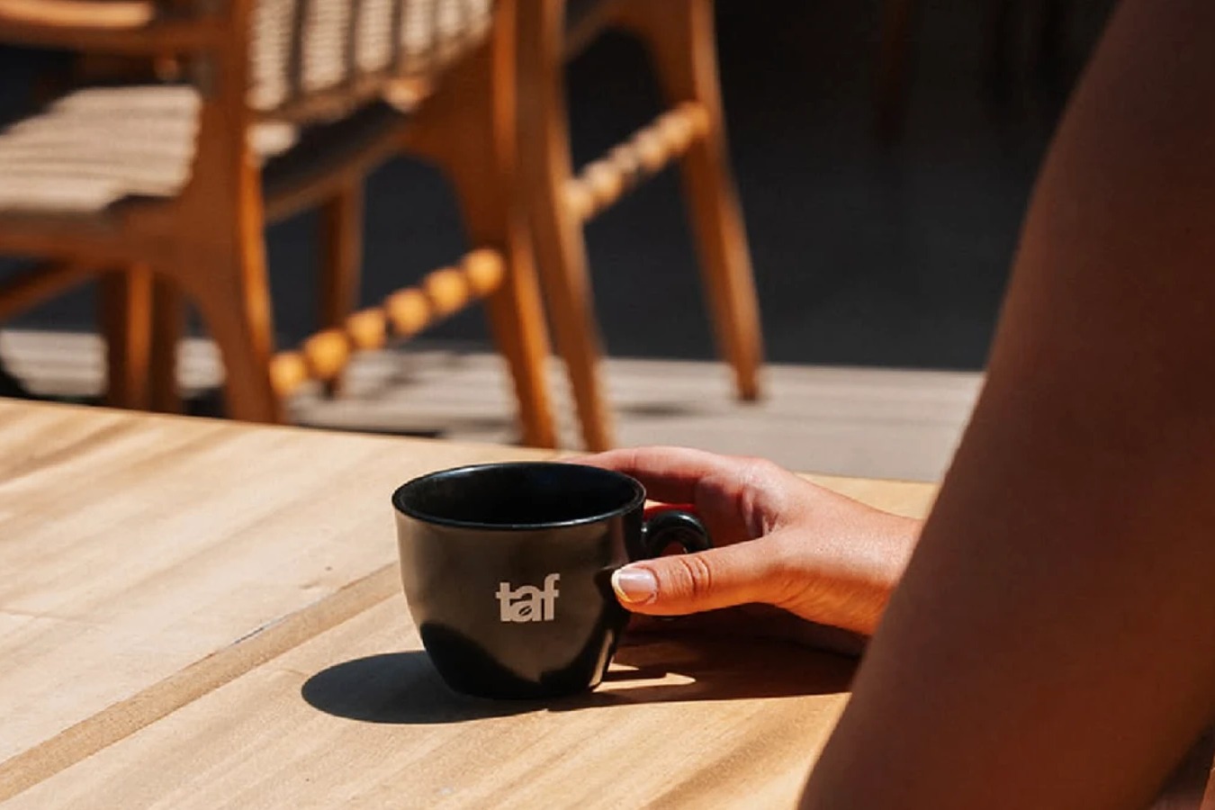 An empty black TAF branded cup resting on a sunlit wooden outdoor table, wicker chairs in the background