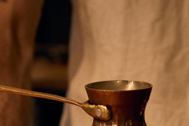 A hammered copper cezwe coffee pot with a long handle held over a cloth backdrop