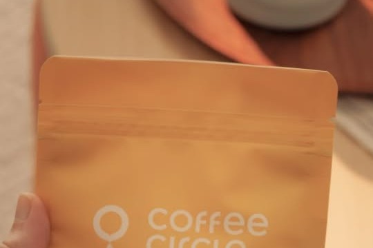 A hand holding a Coffee Circle retail coffee bag with a cup of black coffee blurred in the background