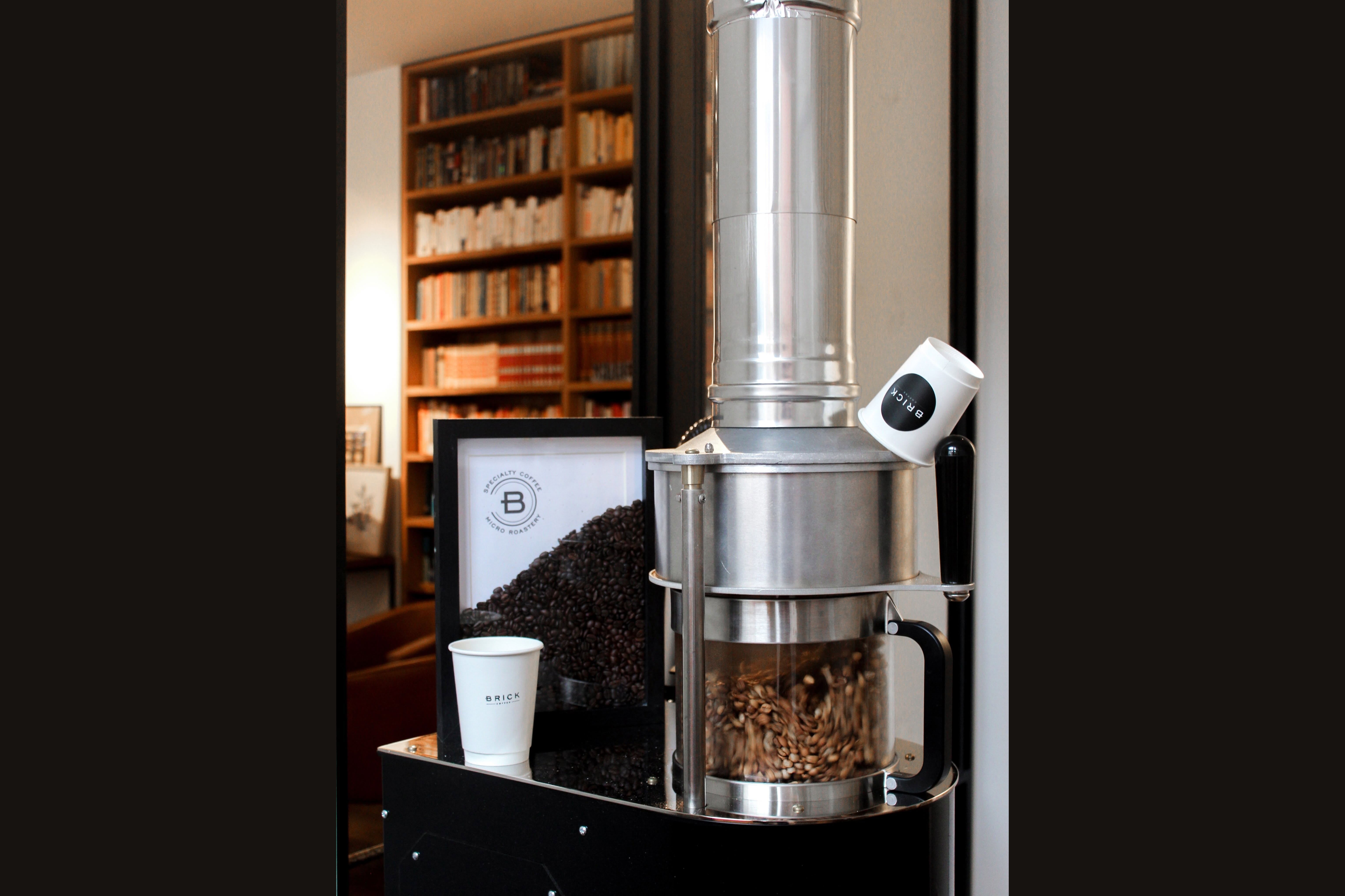 The on site roaster with a hopper of green beans, a framed Brick Coffee micro roastery poster, and a branded cup on the cart