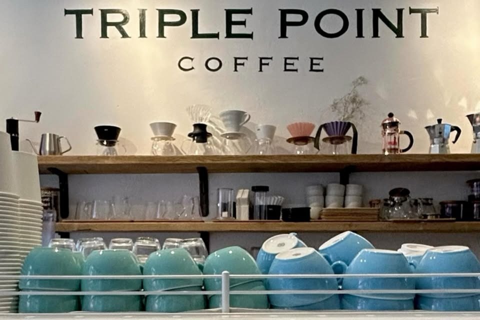 The TRIPLE POINT COFFEE wall sign above open wooden shelves stocked with pourover drippers, moka pots, and stacked pastel mugs