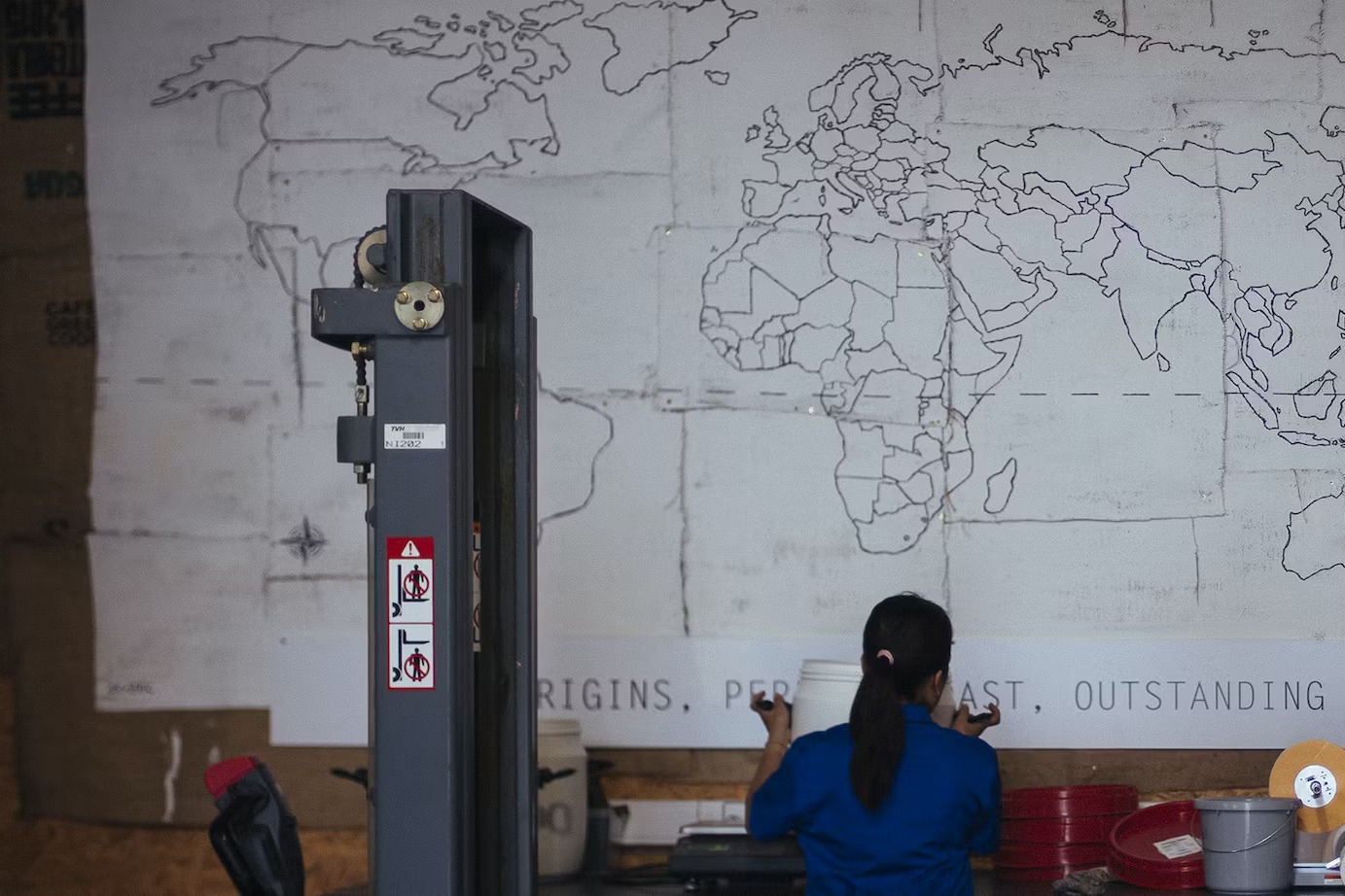 A staff member working at a bench inside OR Coffee's roastery, below a large hand-drawn world map mural reading 'origins, perfect roast, outstanding coffee'