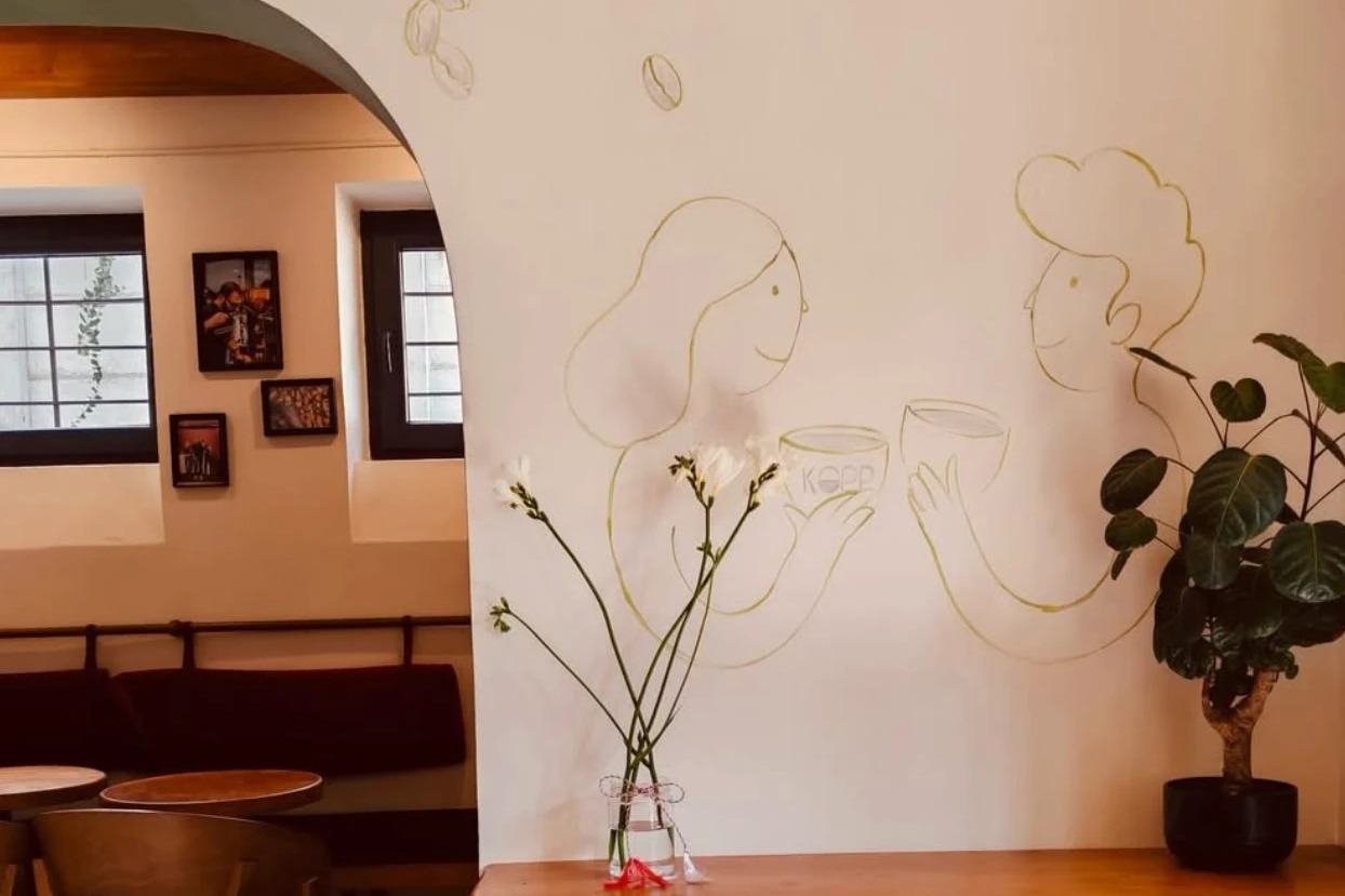 A hand drawn mural of two people toasting KOPP branded cups above a wooden counter with fresh flowers and a potted plant