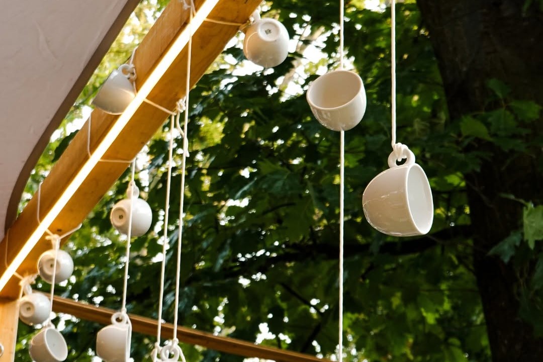 Rows of white teacups suspended from a wooden beam over Origo's outdoor terrace, hanging against a backdrop of green trees