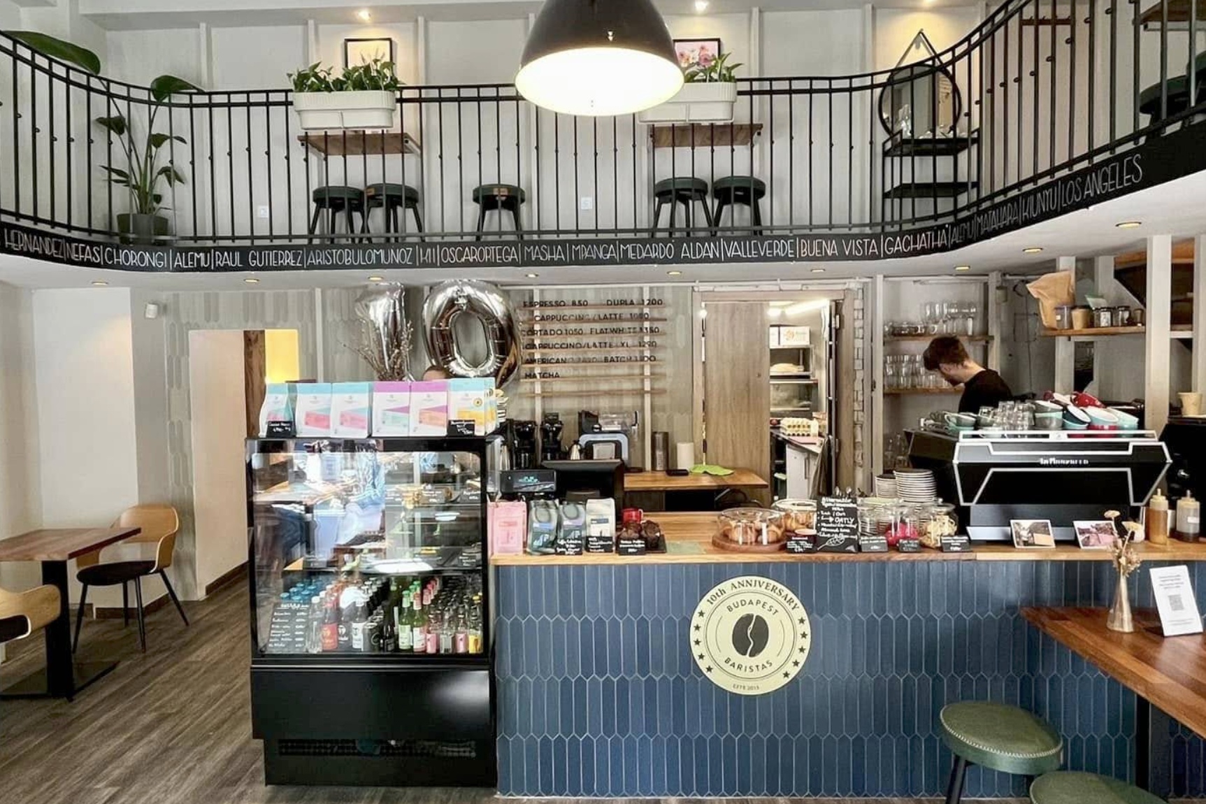 The counter and gallery level at Budapest Baristas, a 10th anniversary logo tiled into the counter front and a chalkboard menu behind the espresso machine