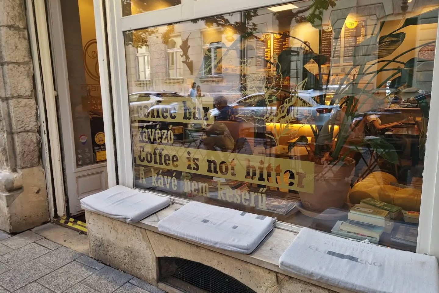 Mantra's front window with its gold lettered Coffee is not bitter, A kávé nem keserű slogan, books and plants visible inside and window bench seating below