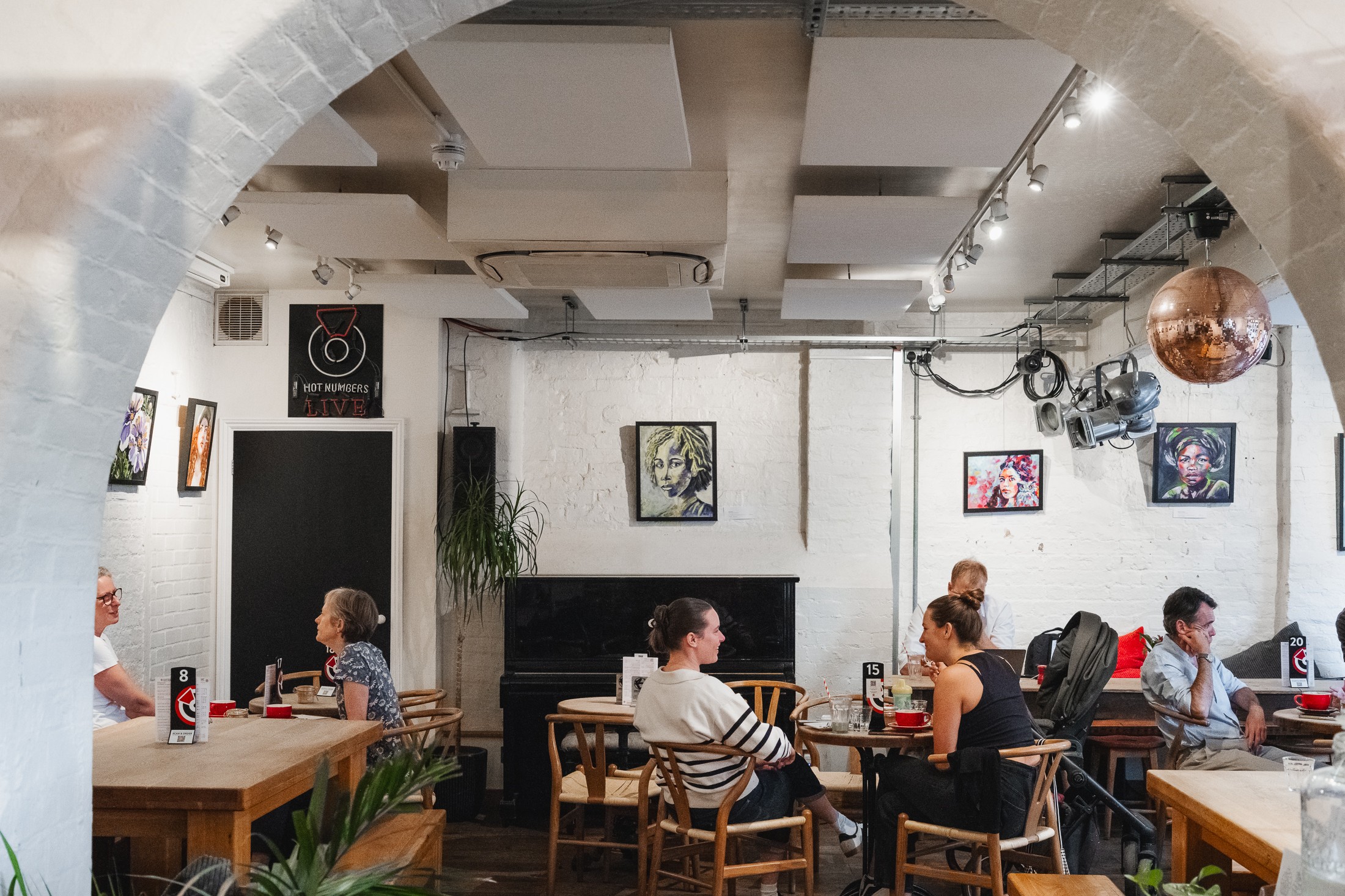 The brick archway interior of Hot Numbers on Gwydir Street, with a neon HOT NUMBERS LIVE sign, portrait artwork on white brick walls, a copper disco ball and communal wooden tables