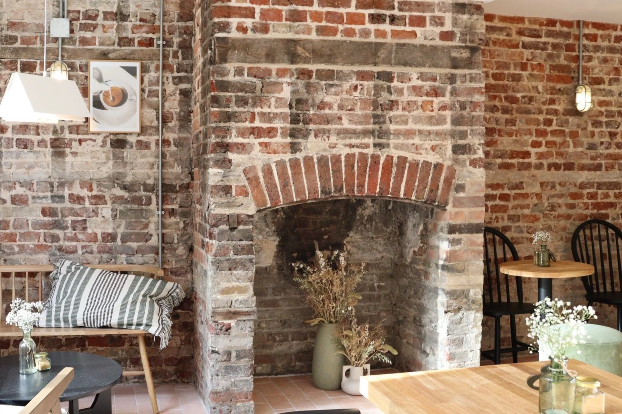 The exposed brick original fireplace inside Pages Coffee House, with dried flowers in ceramic vases, scandi style wooden chairs and a striped cushion on a bench