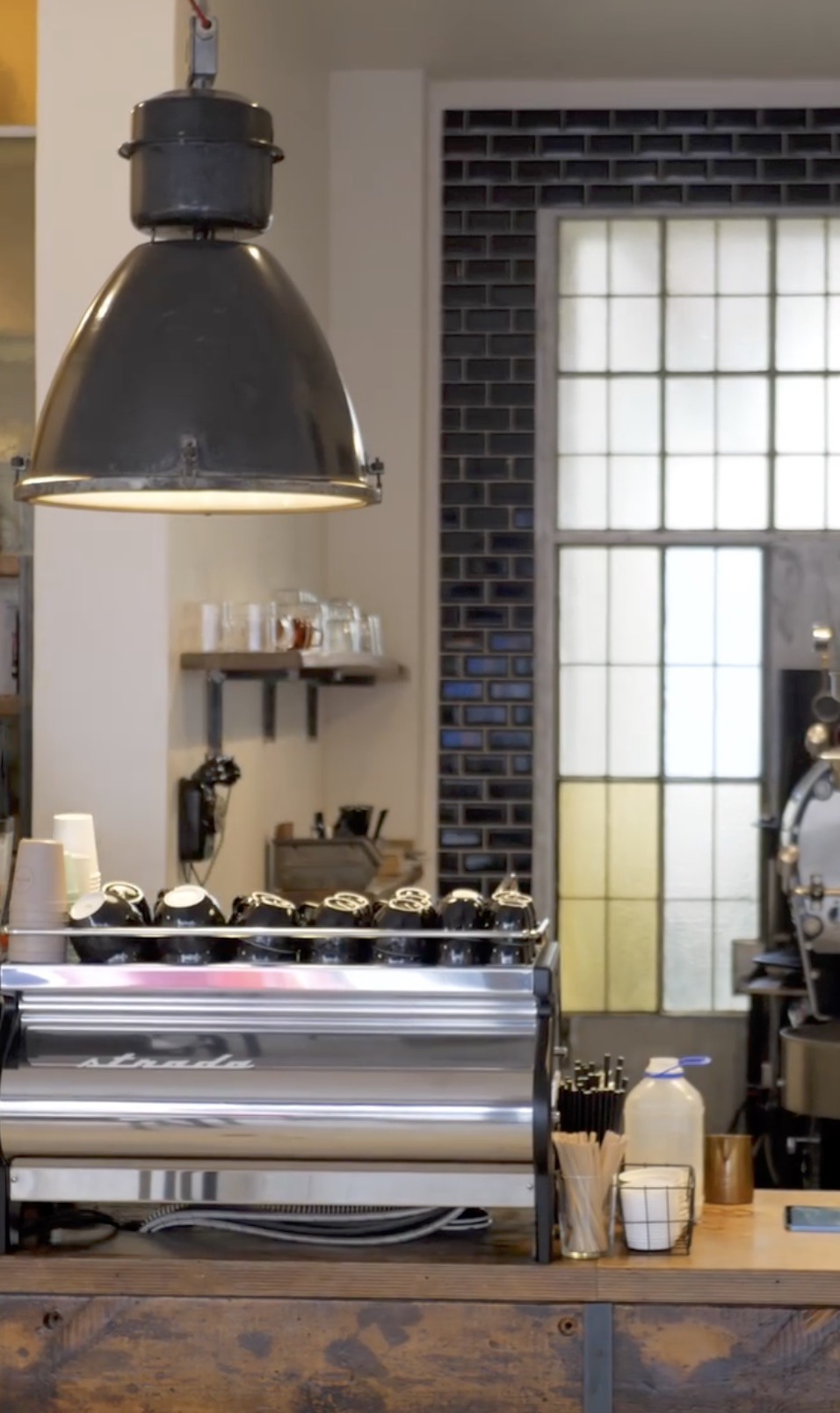 An industrial pendant lamp hanging over the espresso bar, a run of black cups lined up on the machine and a roaster visible through the doorway behind