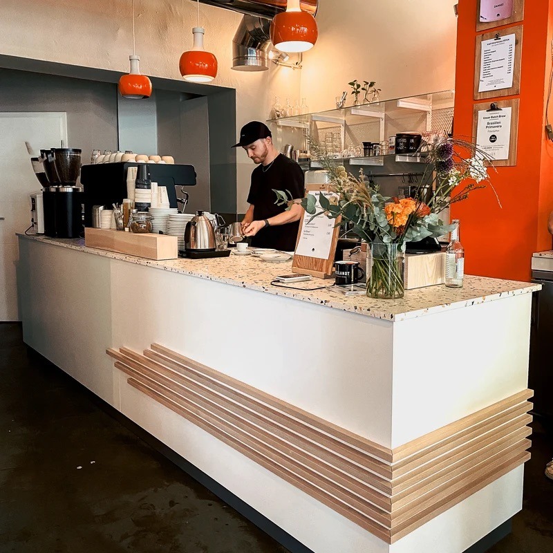 A barista working the terrazzo counter under orange pendant lamps, a grinder and espresso machine to one side and a bouquet of flowers on the counter