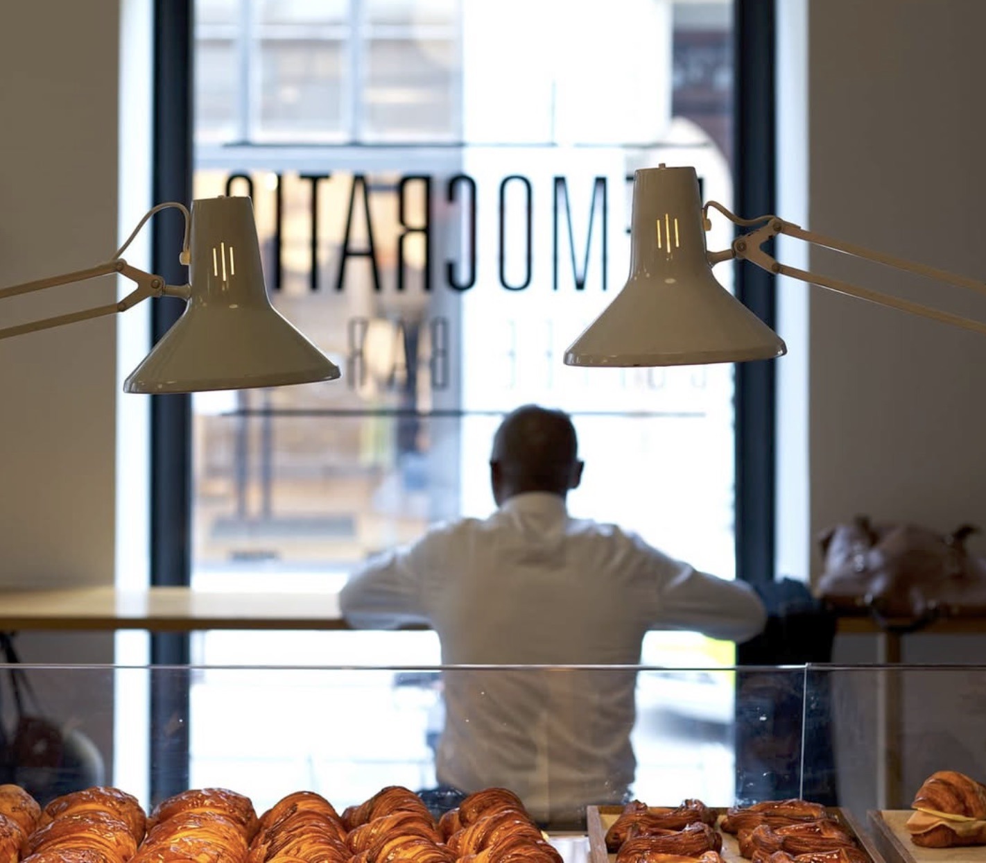 The Democratic Coffee Bar name etched on the front window, a barista silhouetted against the daylight behind a tray of croissants