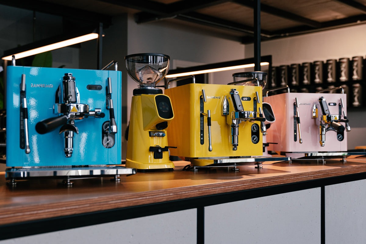 A row of colorful espresso machines and grinders in teal, yellow and pink on a wooden counter, with rows of coffee bag canisters shelved behind