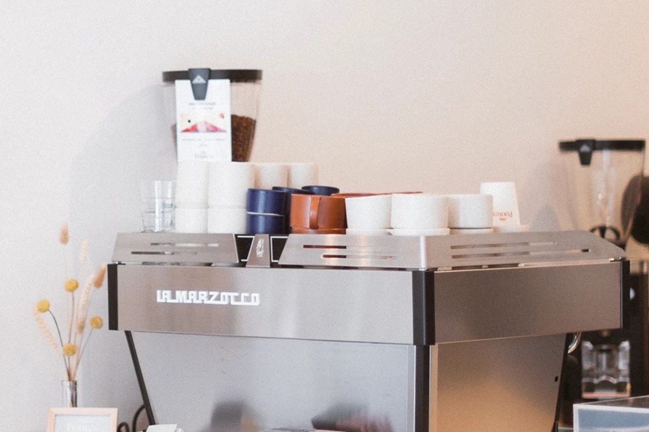 The La Marzocco espresso machine at Pontes, stacked cups and a bag of retail beans on top against a warm plaster wall