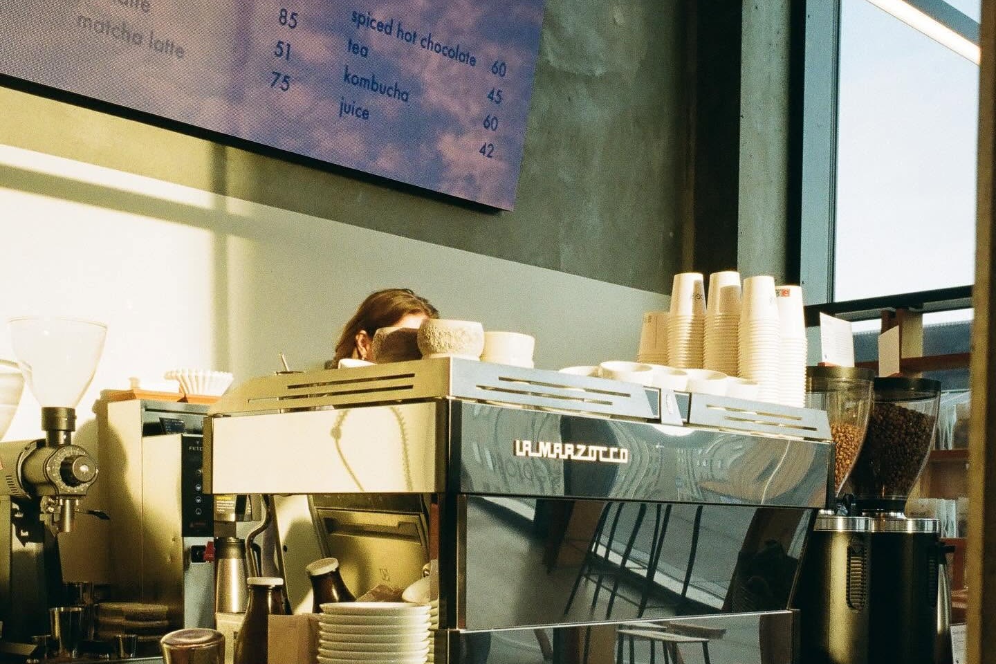 The espresso bar at Prolog Coffee Bar with a La Marzocco machine, stacked cups, and a blue menu board reading batch brew, pour over, and matcha latte