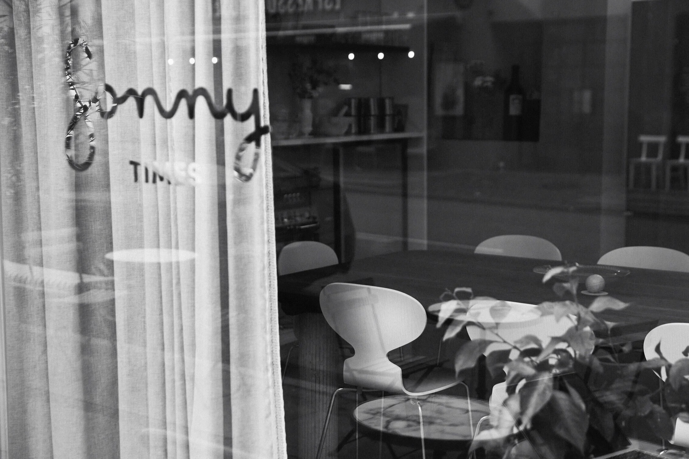 Black and white view through Sonny's curtained storefront window, showing Fritz Hansen Ant chairs and a marble cafe table inside