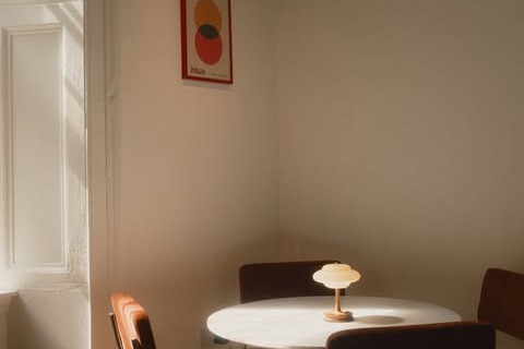 A sunlit corner inside Bono with a round white table, orange upholstered chairs, a small table lamp, and a red framed print on a pale wall