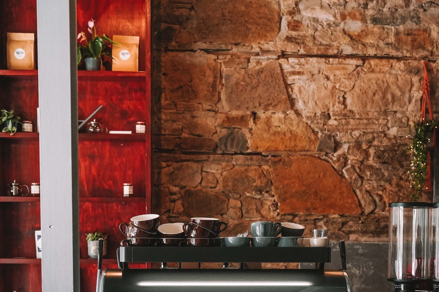 Tanifiki's counter with a black Dalla Corte espresso machine in front of exposed stone wall and red wooden shelving stocked with Rafiki branded coffee bags and plants