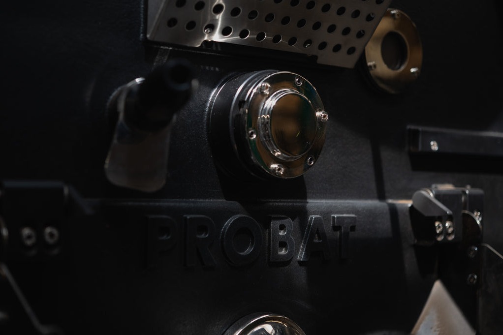 Close-up of the black Probat roaster's nameplate with PROBAT embossed in the metal casing and polished chrome fittings catching the light