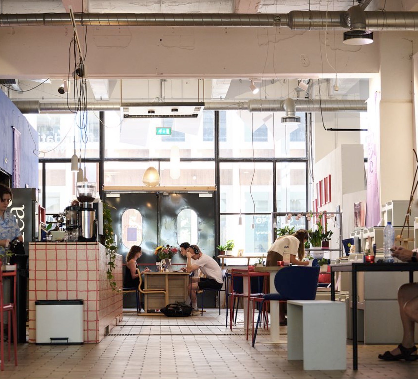 The high ceilinged room at Community Square, a red gridded tile counter to one side and people working at communal tables under exposed ductwork