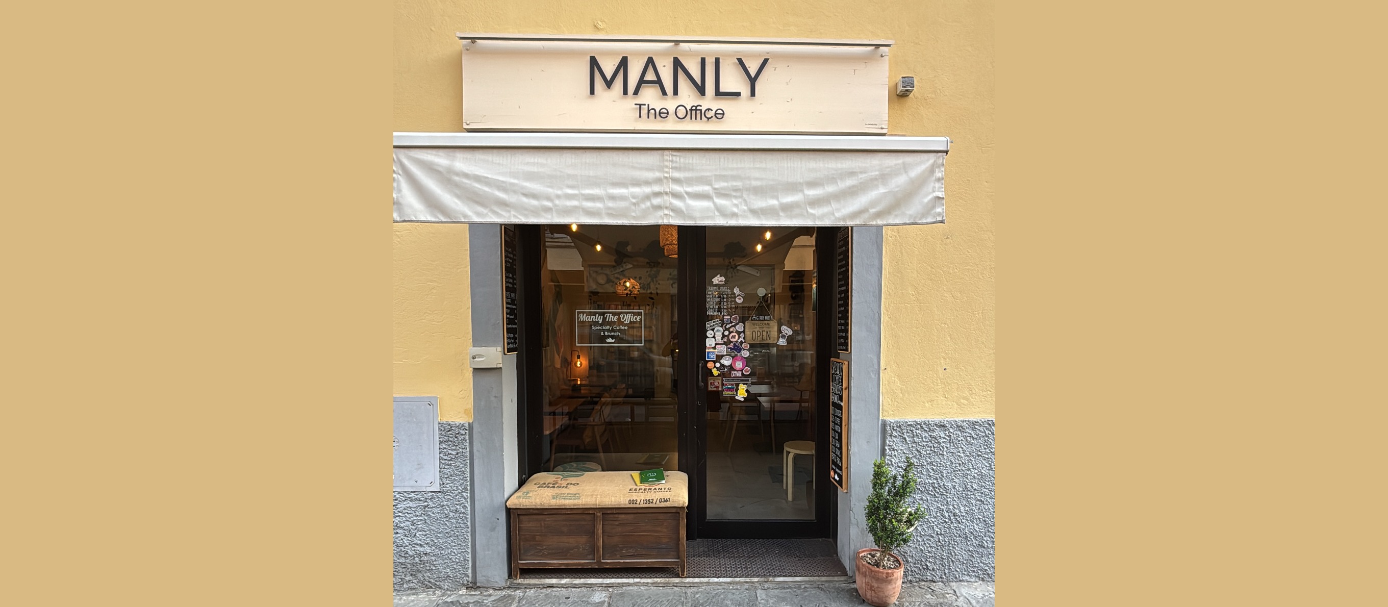 The MANLY The Office sign above the full glass entrance, tables and hanging lights visible inside, a burlap sack bench and potted plant out front