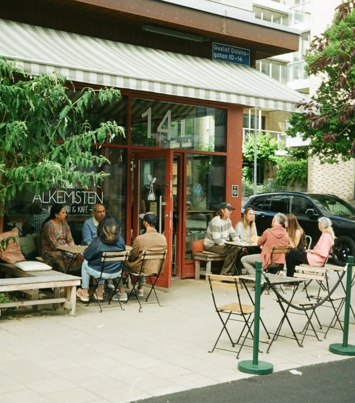 The striped awning and red framed entrance of Alkemisten on Gustaf Dalénsgatan, with customers seated at outdoor tables