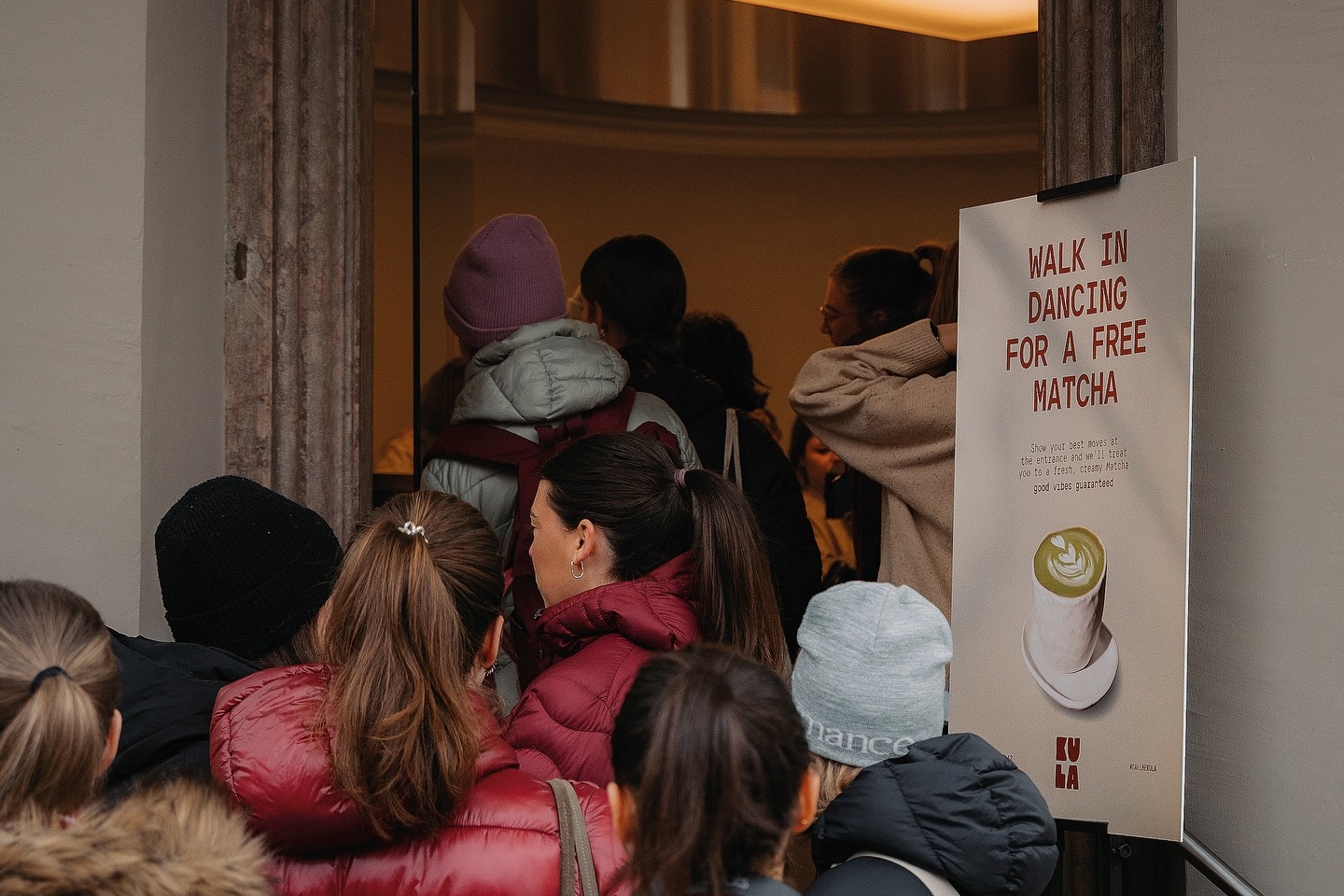 A line of customers, seen from behind, queuing at Kula.Lab's entrance beside a sign offering a free matcha for a dance move, warm light spilling from the doorway
