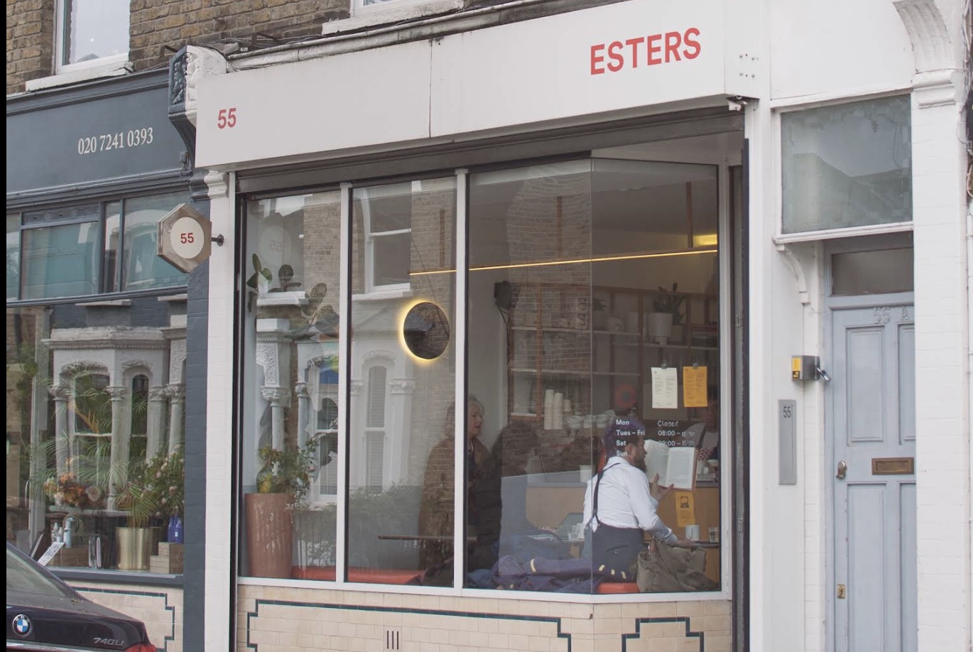 The Esters storefront at 55 Kynaston Road, a customer seated inside reading beneath a round pendant light and the shop's posted hours