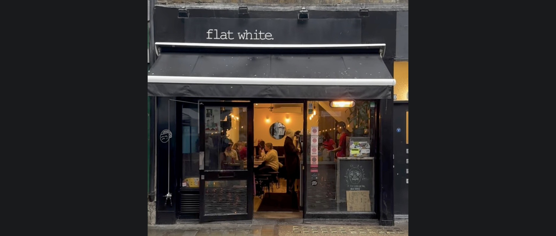 The 'flat white.' signage and awning above the open storefront, with customers seated inside under warm lighting