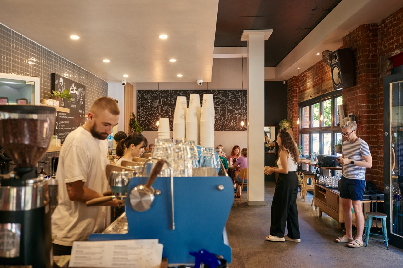 A barista pulling shots at the blue espresso machine while customers order at the counter inside the exposed brick Collingwood warehouse