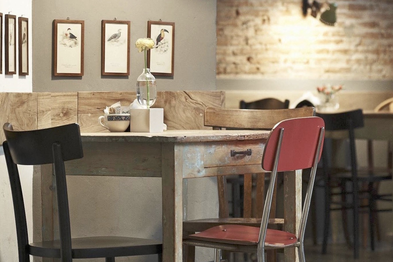A vintage wooden table set against exposed brick at Nicol, framed bird prints on the wall and a mix of black and red chairs