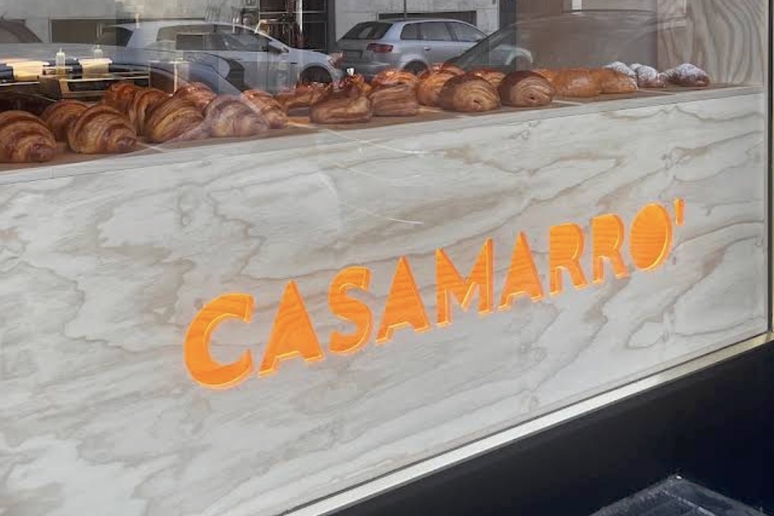 The CASAMARRO' wordmark painted on the storefront window, croissants and pastries lined up in the case behind the glass