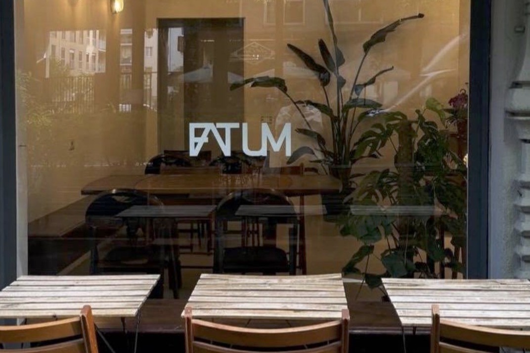 The FATUM wordmark etched on the window above outdoor tables, with wooden interior tables and large potted plants visible inside