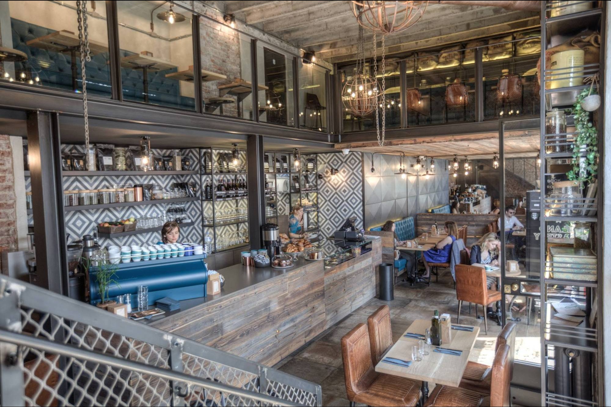 The two level Milano Roastery interior, a cobalt blue espresso machine at the counter beneath an open mezzanine with tufted booths and hanging globe lights