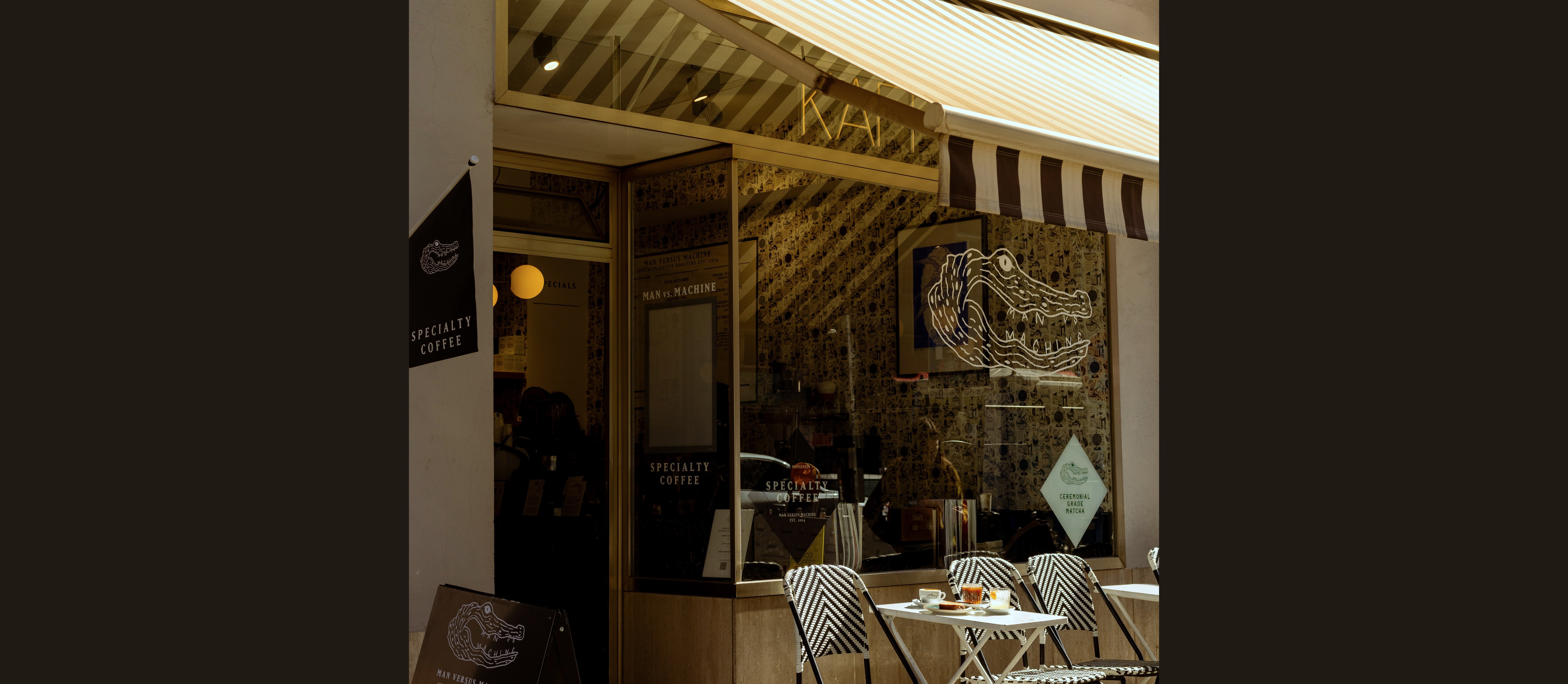 The full Man Versus Machine storefront corner with the striped awning, sandwich board, crocodile logo, Specialty Coffee signage, Ceremonial Grade Matcha diamond sign, and outdoor seating with drinks on the table