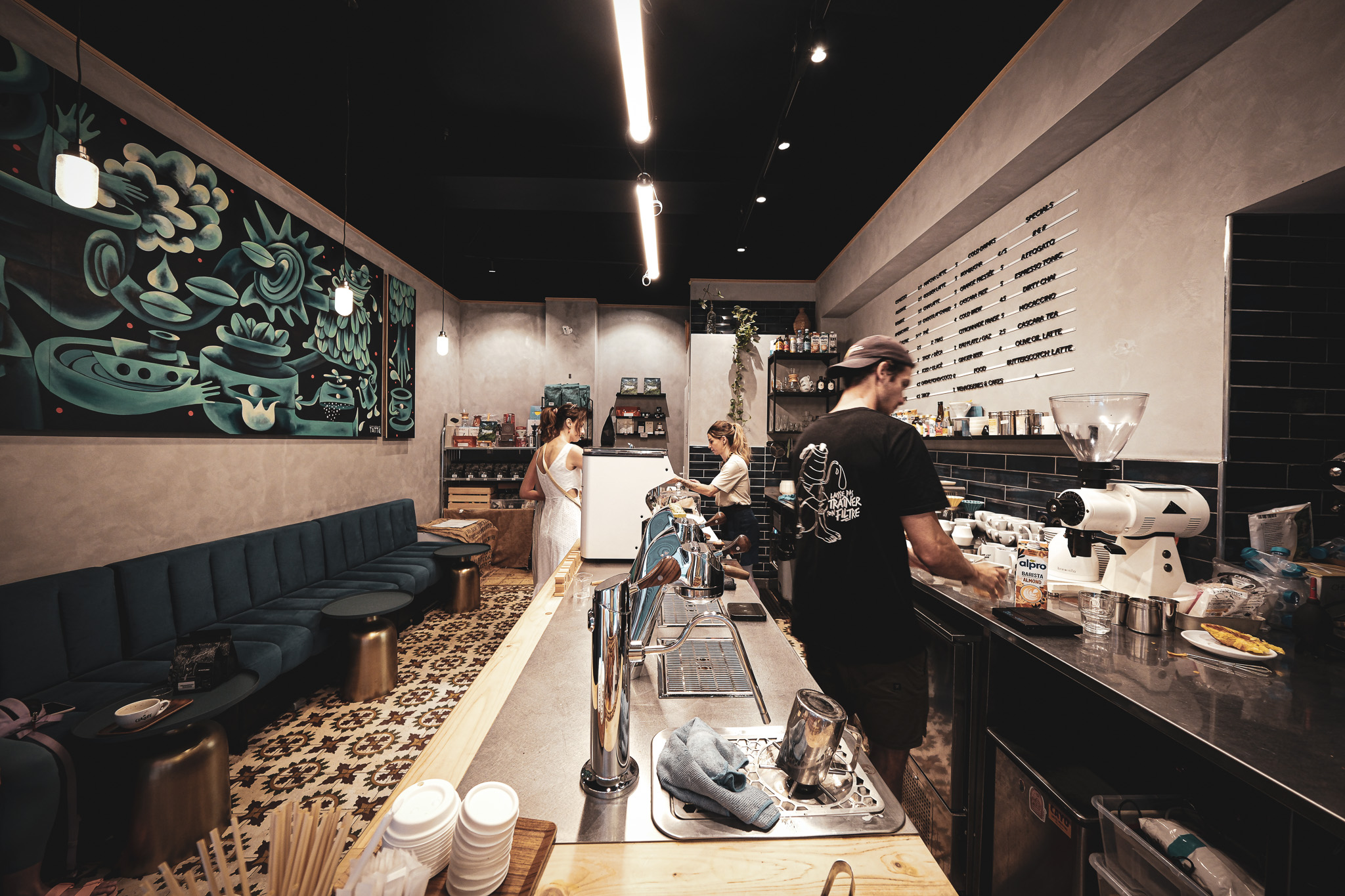 The tiled counter at La Claque Café with baristas working the espresso machine beneath a large mural and a menu board on the wall