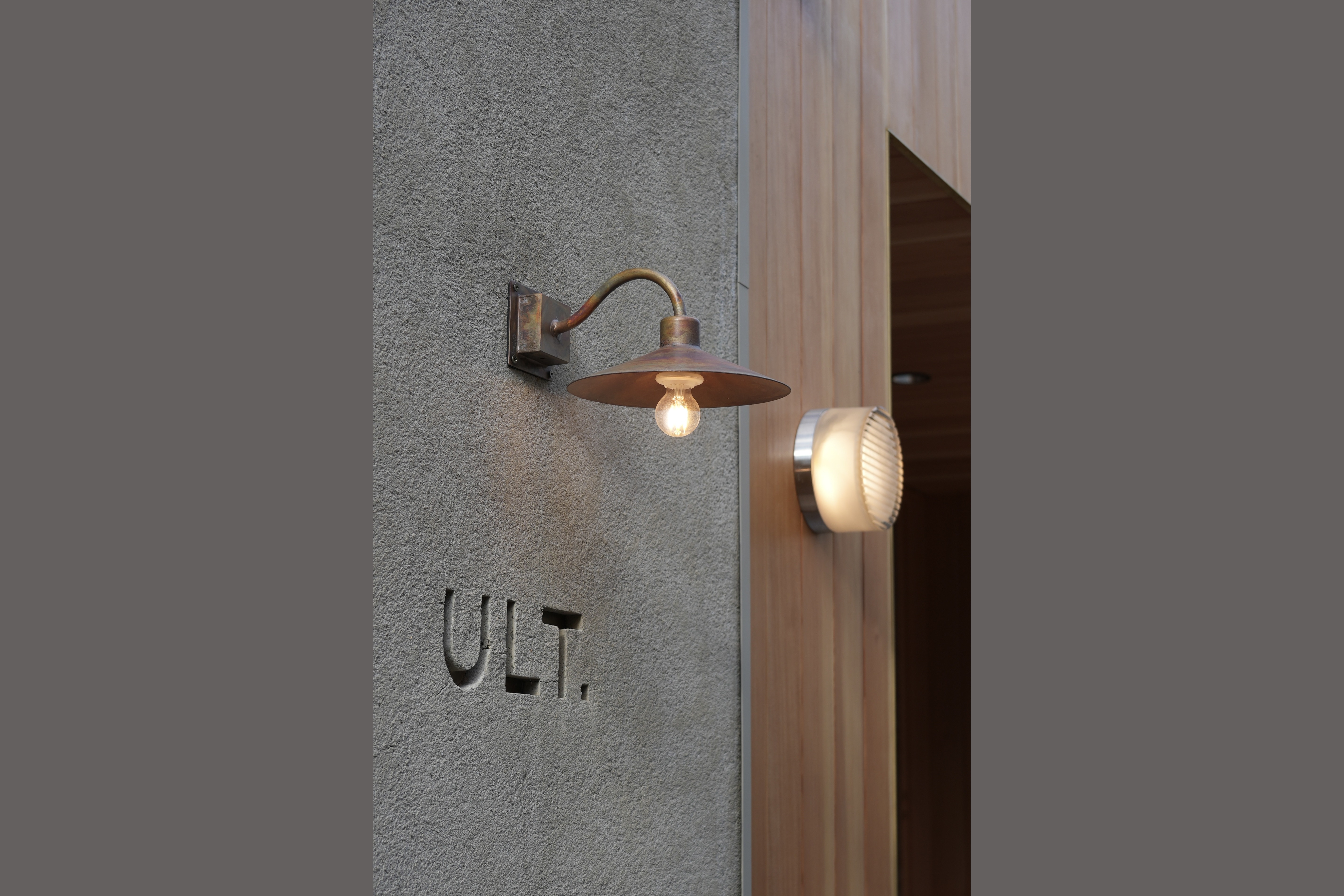 The ULT. lettering carved into the concrete entrance wall beside a brass wall lamp and the wood door frame