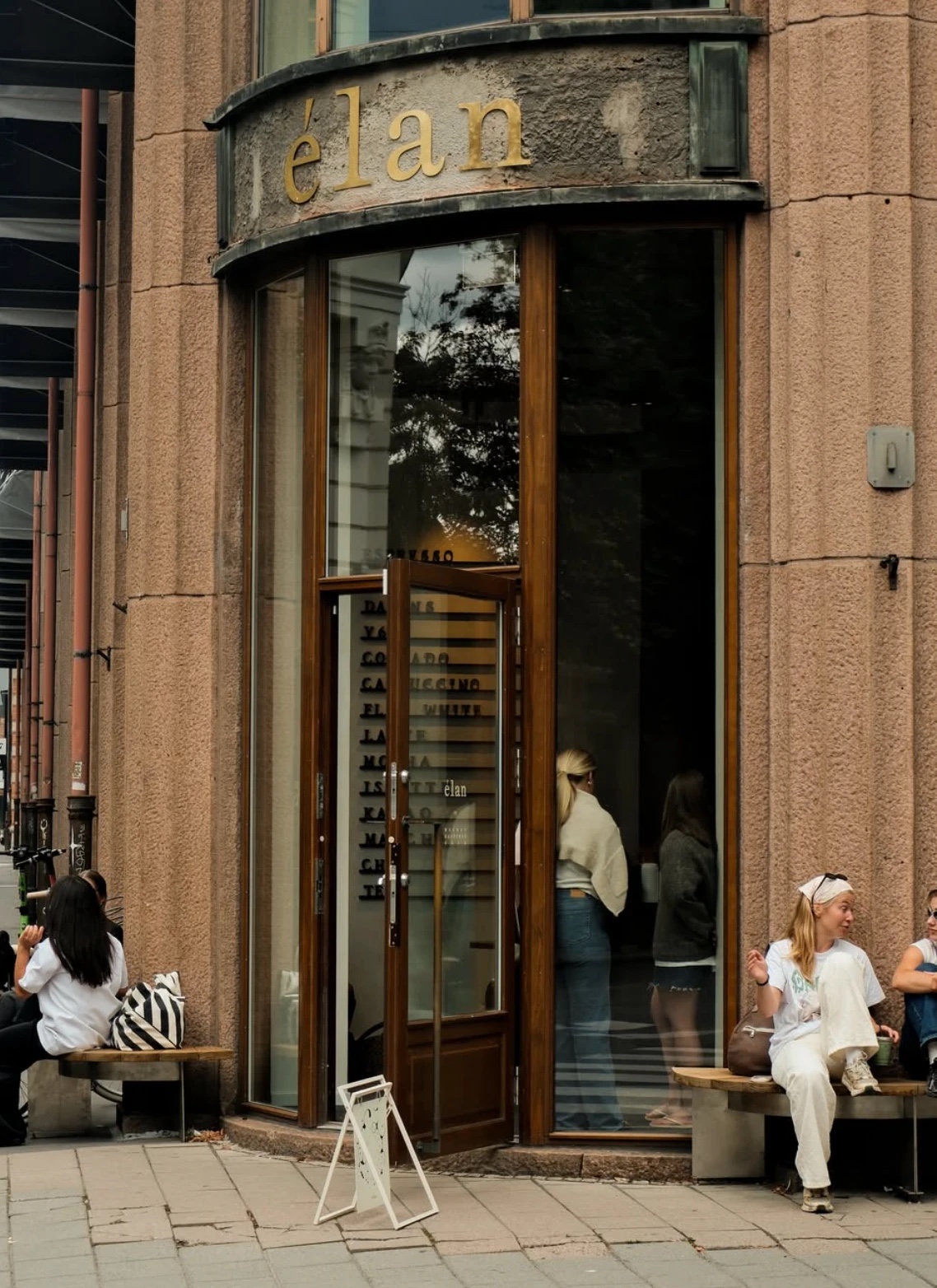 The curved corner entrance of élan, gold lettering on the stone facade of the Sjøfartsbygningen, a drinks list in the window and people sitting on benches either side of the door