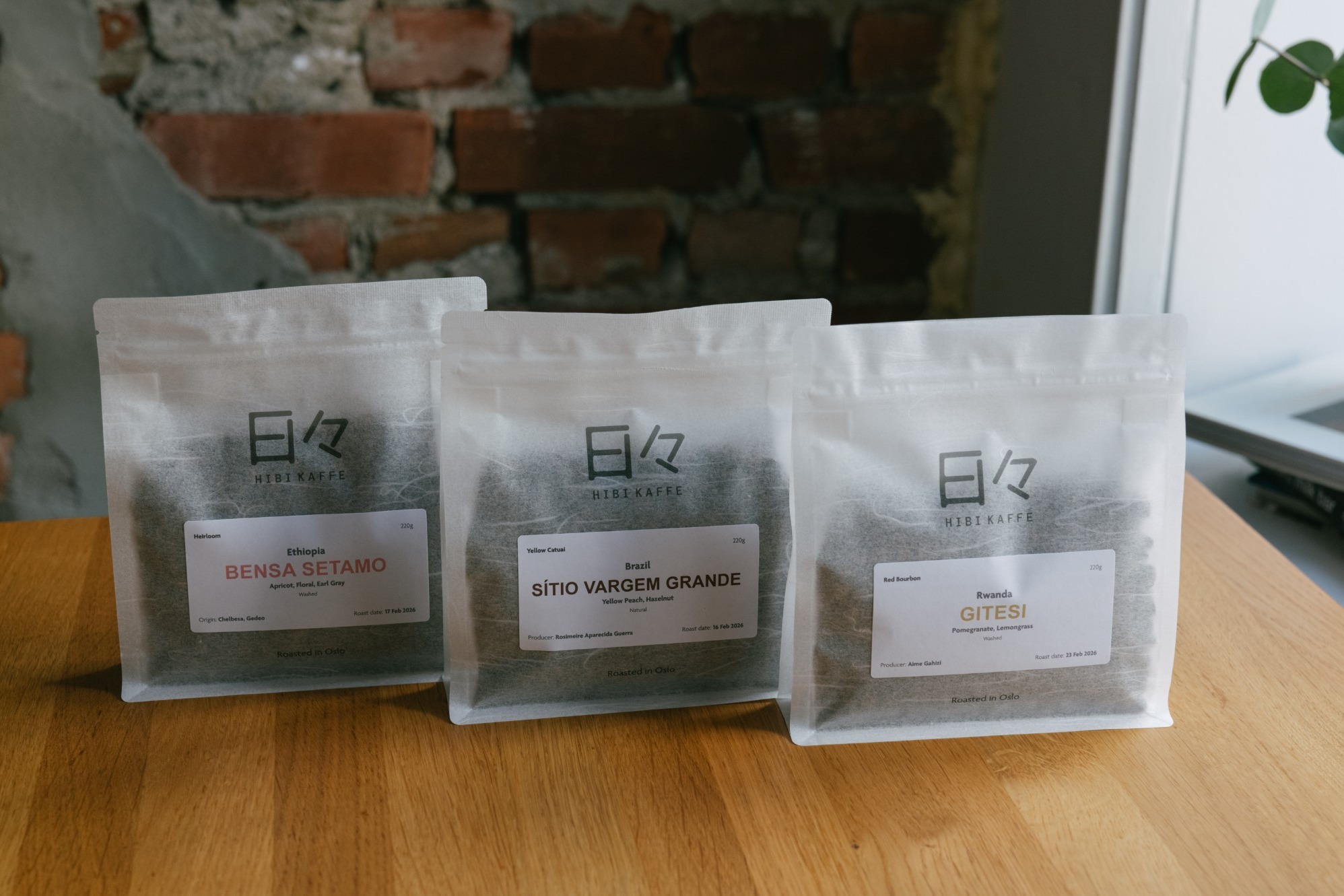 Three Hibi Kaffe coffee bags, Ethiopia Bensa Setamo, Brazil Sitio Vargem Grande, and Rwanda Gitesi, lined up on a wooden table against an exposed brick wall