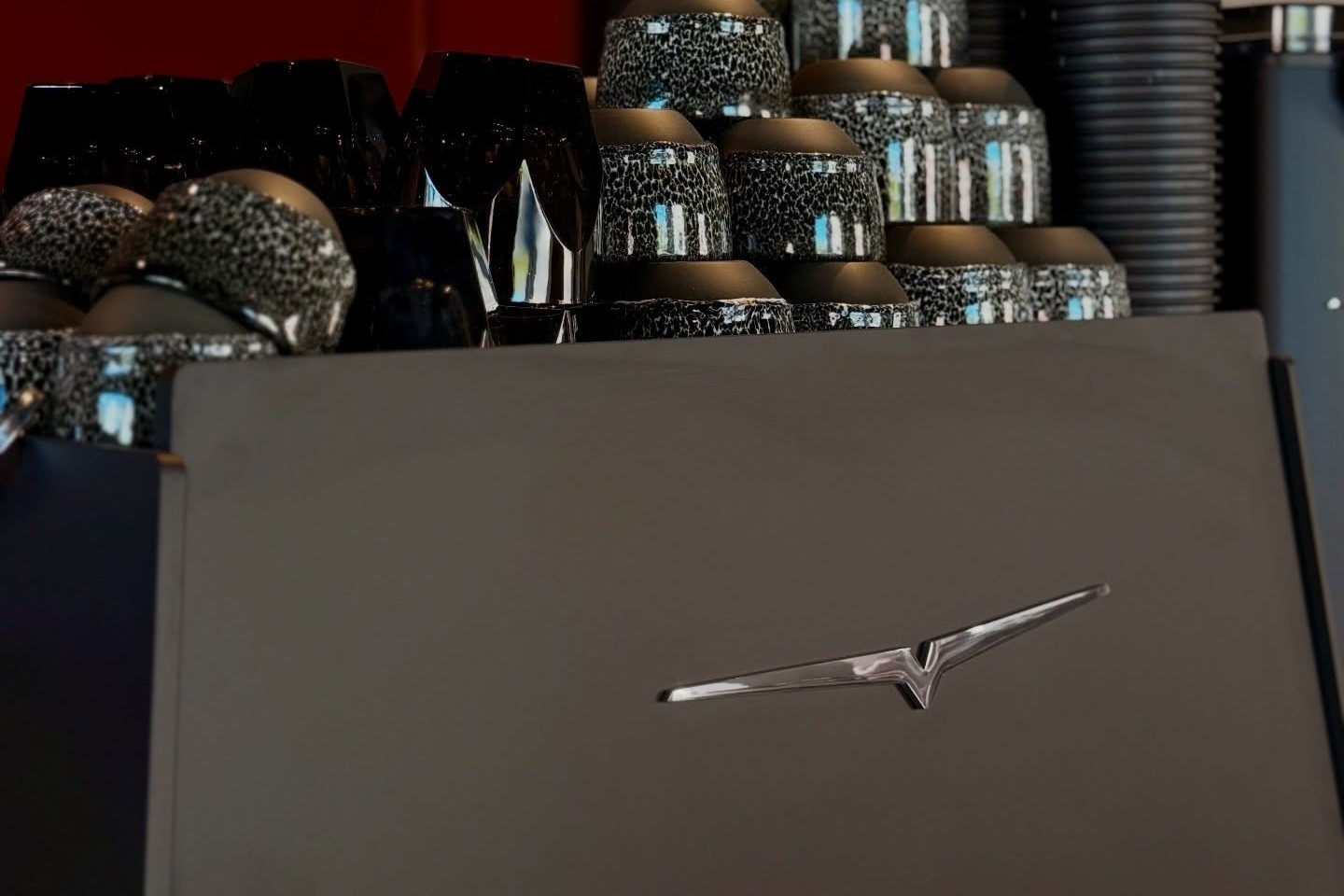Stacks of Mad Drop's speckled ceramic cups and black to go cups behind the counter, with the Victoria Arduino espresso machine's chrome badge in the foreground