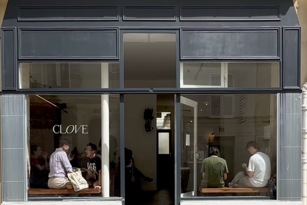 The Clove storefront on Rue Chappe, grey blue paneling and tiled pilasters with the CLOVE wordmark etched on the window and customers seated on the bench inside