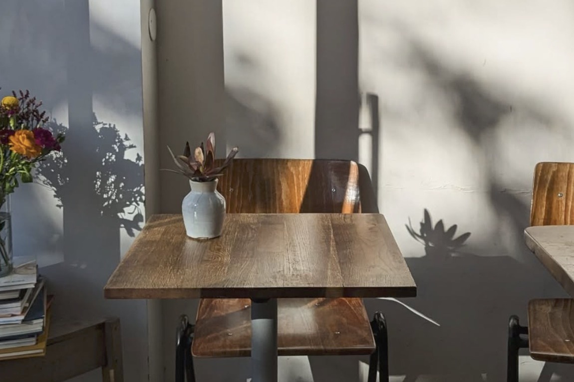 A sunlit table at I.O Café with a small stoneware vase of dried flowers, wooden bentwood chairs and dappled shadow on the wall
