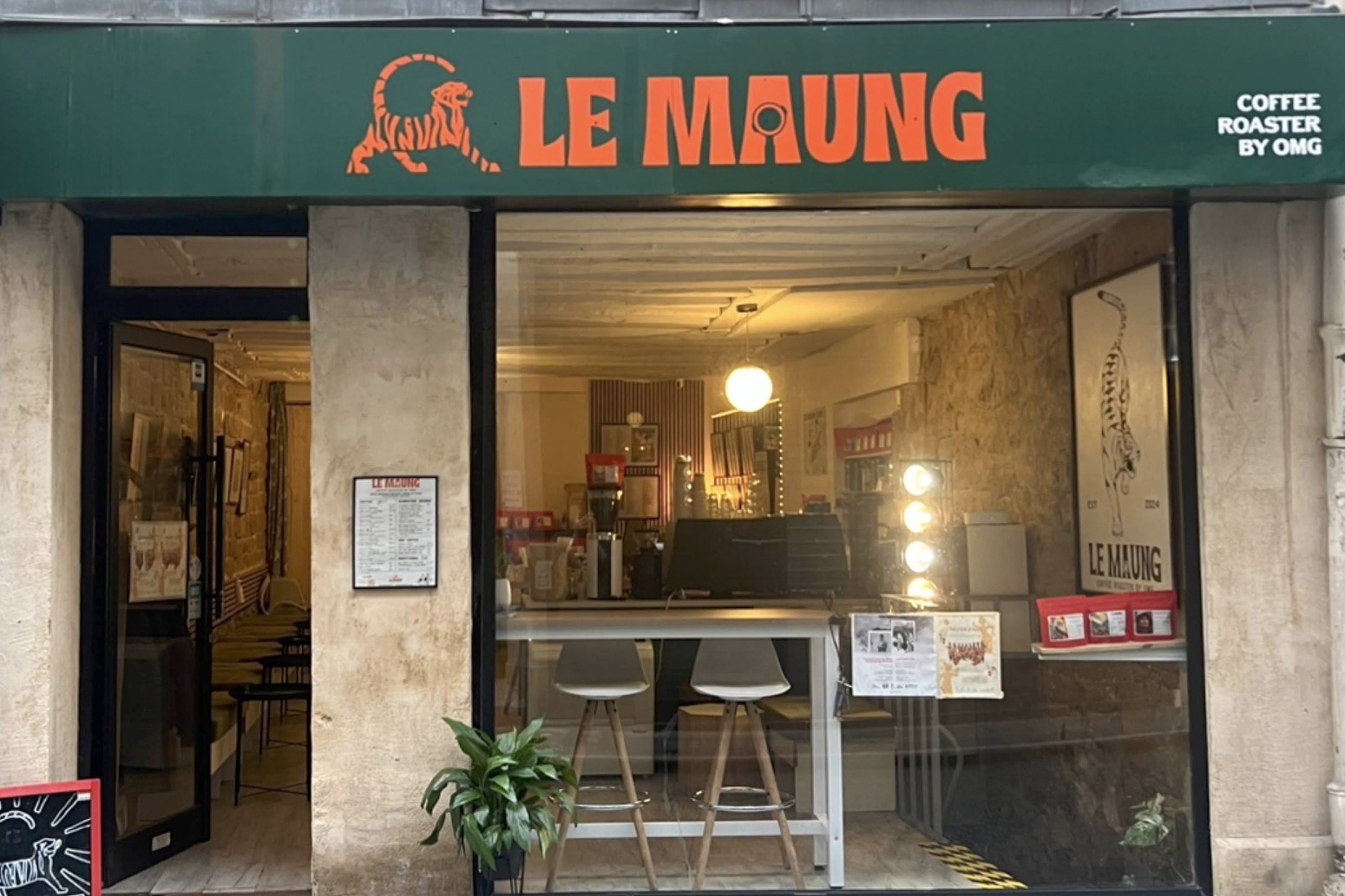 The green Le Maung storefront with its orange tiger logo, plants and bar stools at the counter visible through the open window