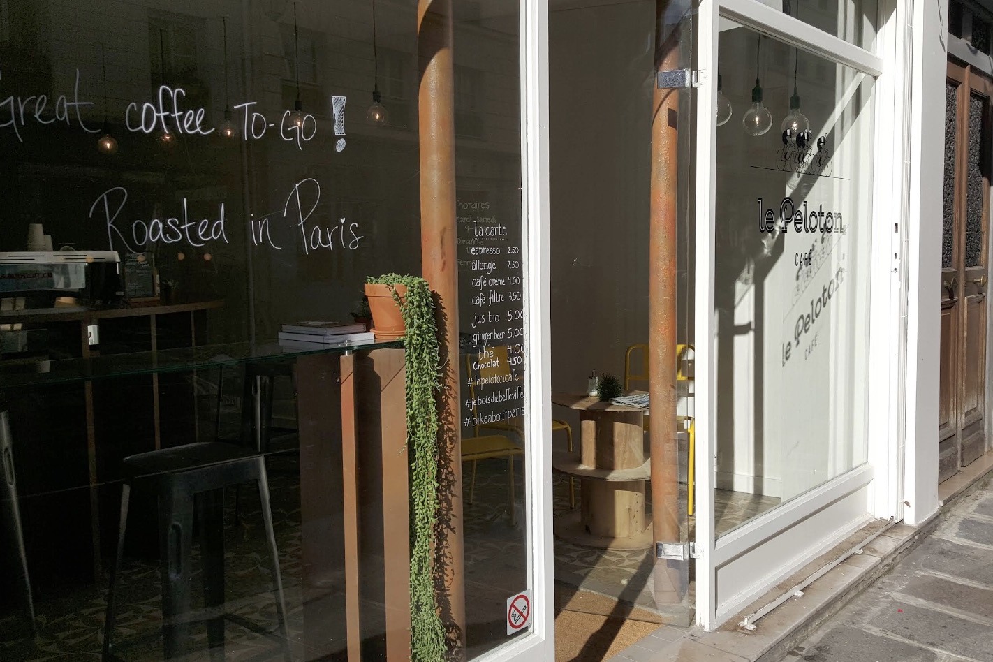 The Le Peloton Café window, hand chalked Great coffee to-go and Roasted in Paris beside the menu board and the le Peloton sign through the open door