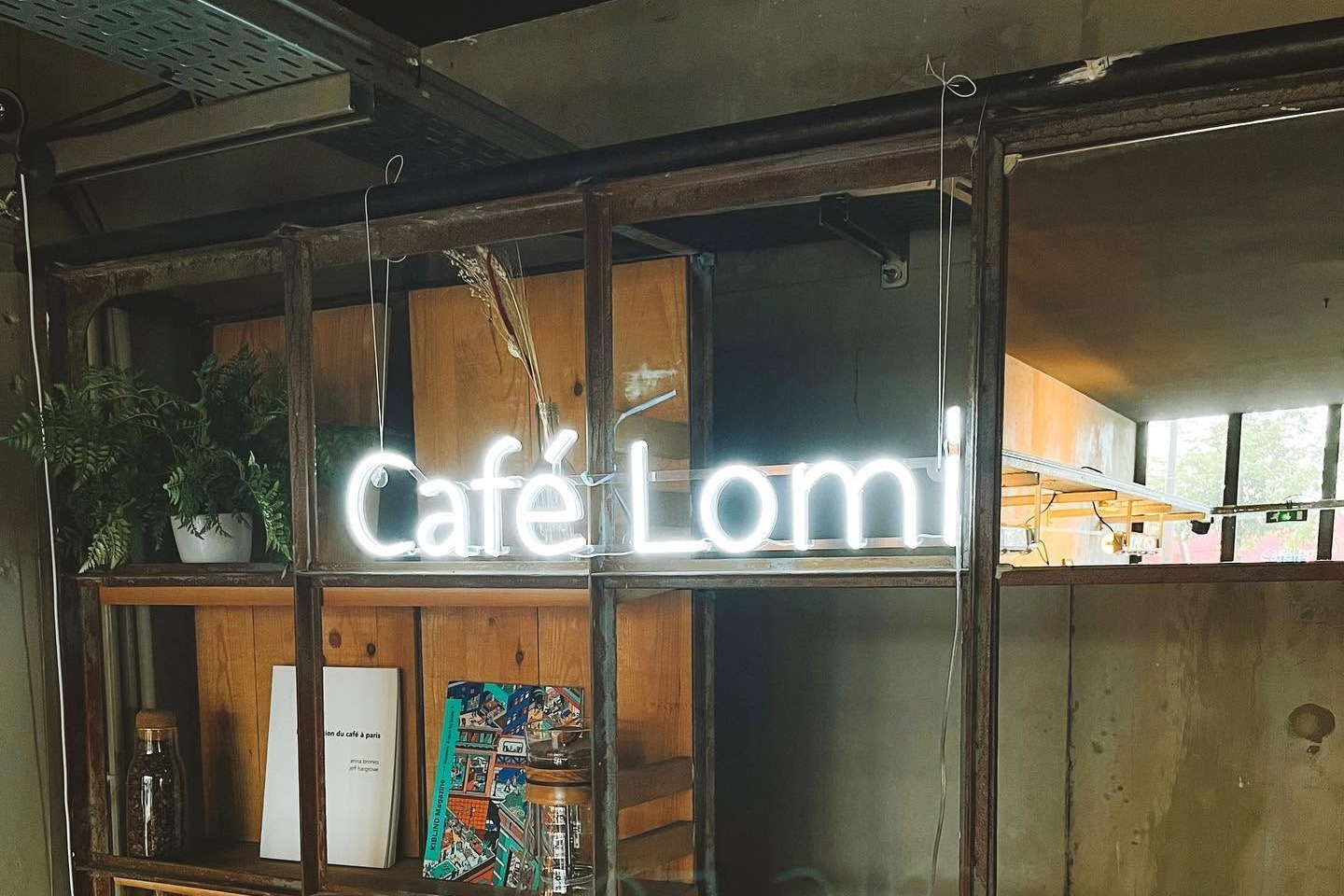 The Café Lomi neon sign glowing above an industrial shelving unit with plants, books and roasting ephemera