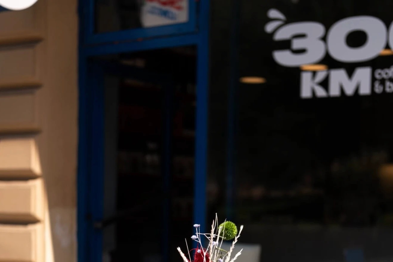 The 300 KM coffee and bakery window sign seen past a dried flower arrangement on an outdoor bistro table