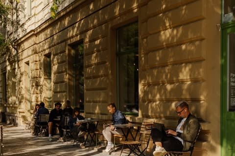 People working on laptops at outdoor tables along the sunlit stone facade outside Muj salek kavy in Karlin