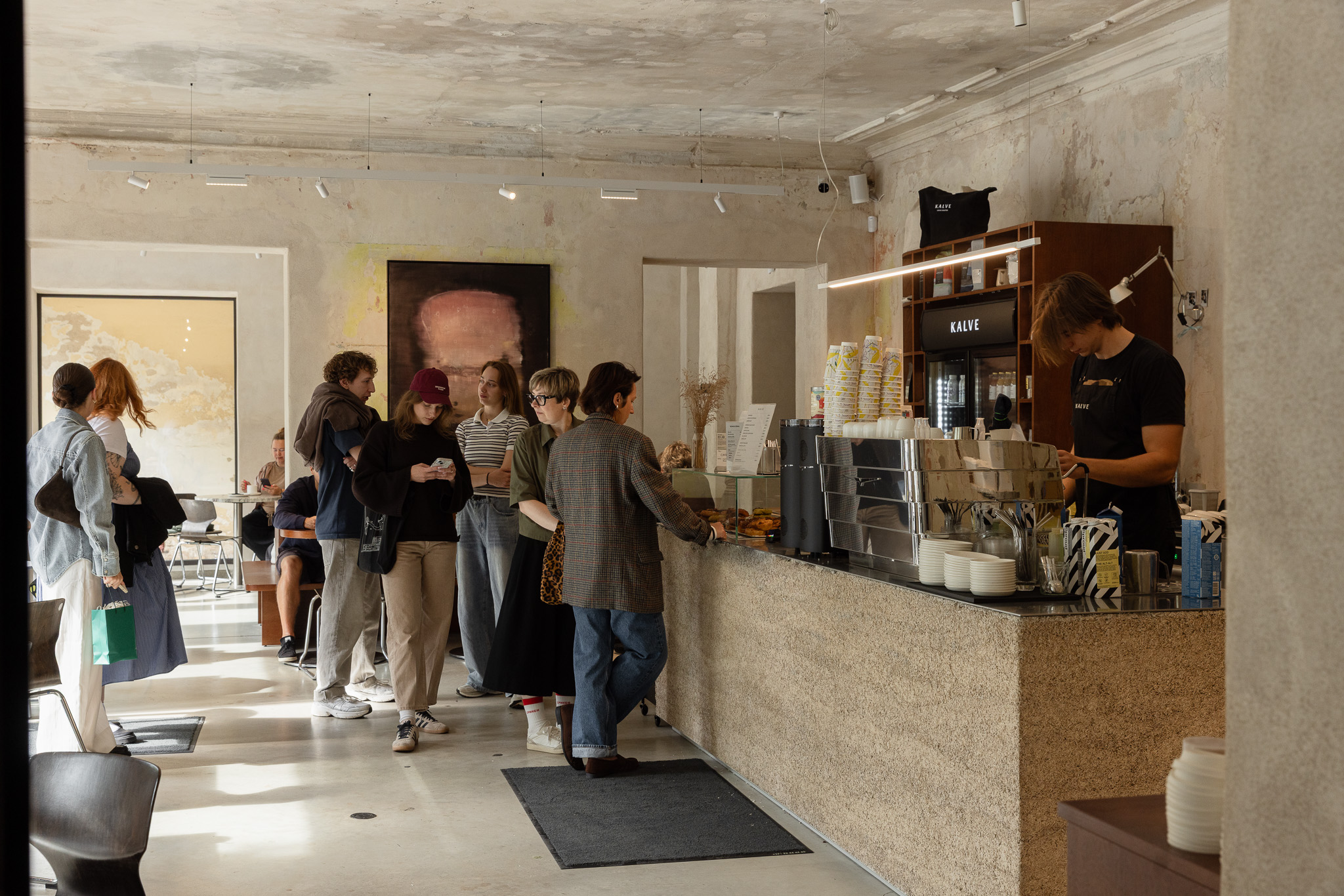 A queue of customers at the KALVE Living Room stone counter, staff working the espresso machine beneath raw plastered walls and an abstract painting